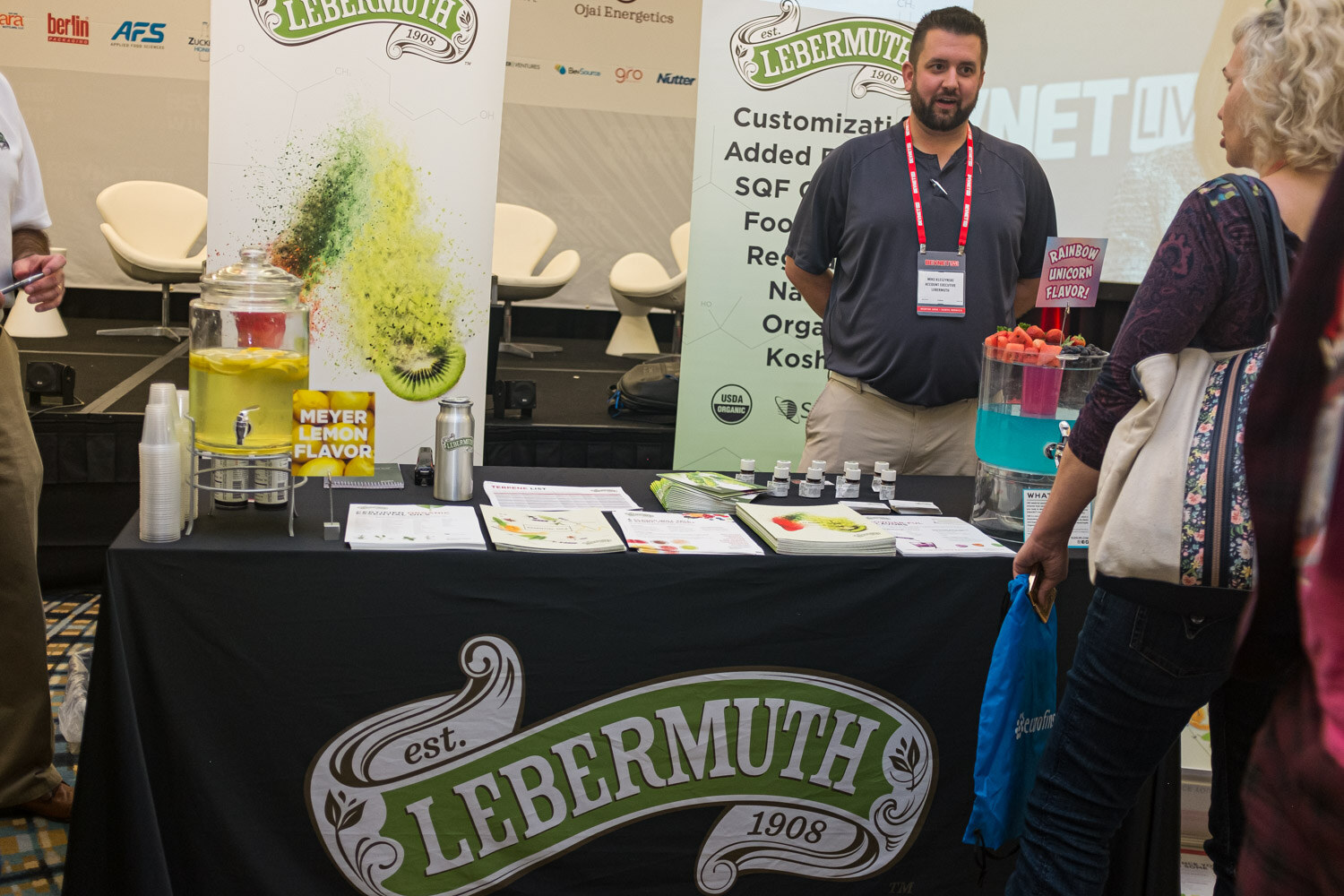 A Lebermuth exhibitor stands behind a black-draped table with an infused-water dispenser and sample cups as an attendee passes by.