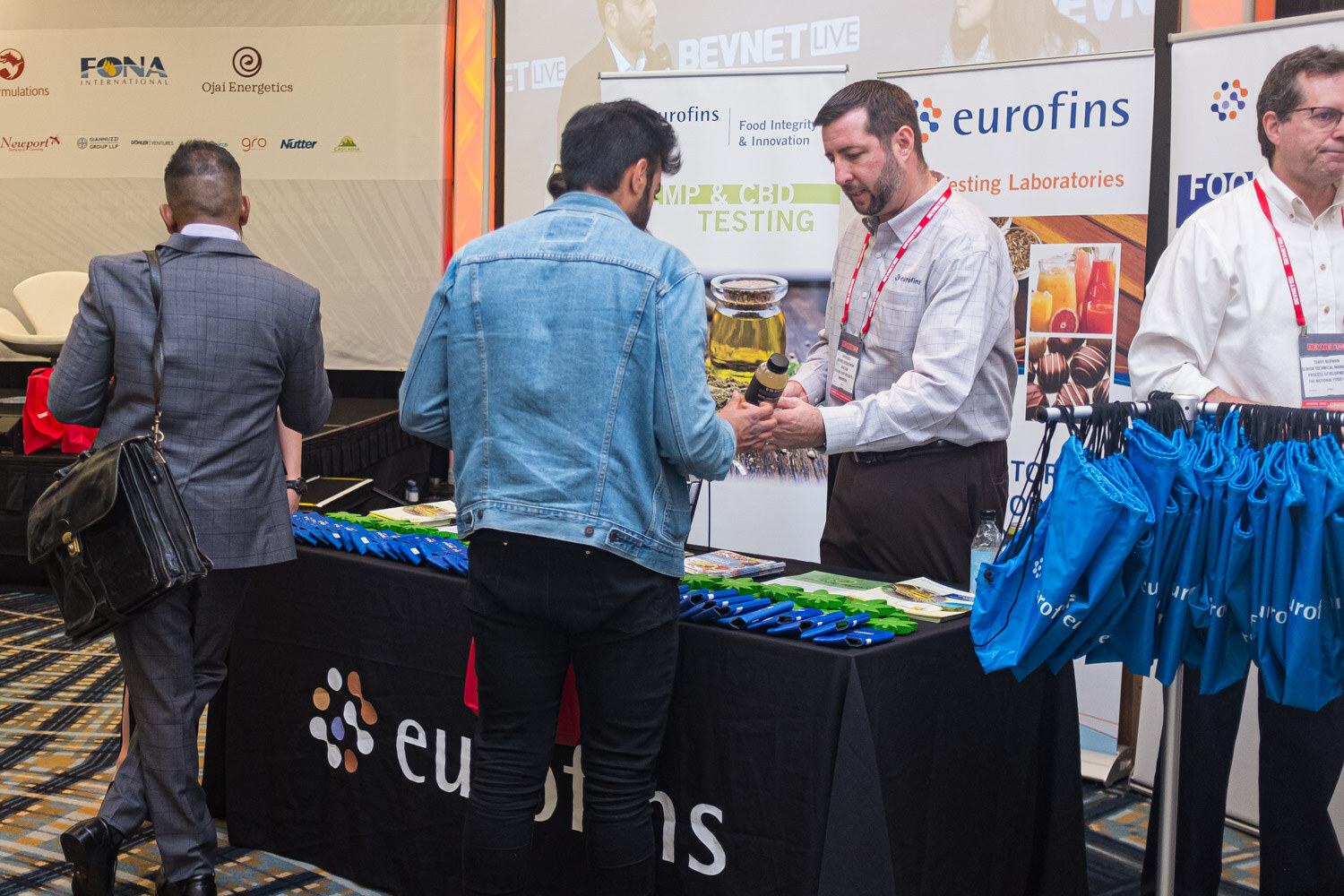 Two attendees and two exhibitors in white shirts stand at a table with eurofins signage and blue tote bags.