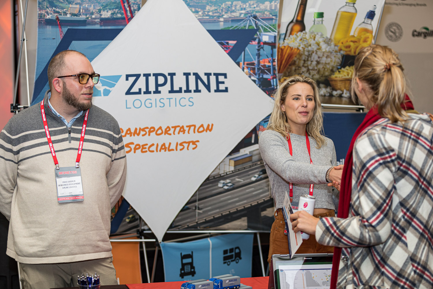 A man with glasses and a woman in a grey sweater stand under a Zipline Logistics diamond sign while a visitor in plaid listens.