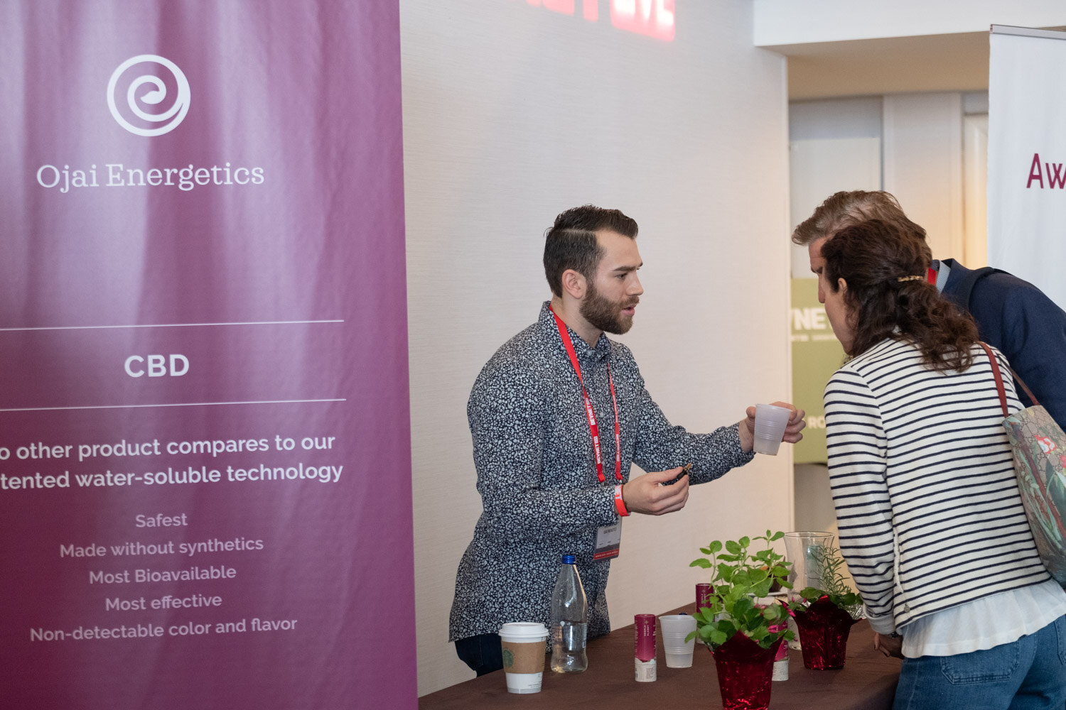 A bearded man in a patterned shirt offers a small cup to a woman in a striped shirt in front of a purple Ojai Energetics CBD banner.