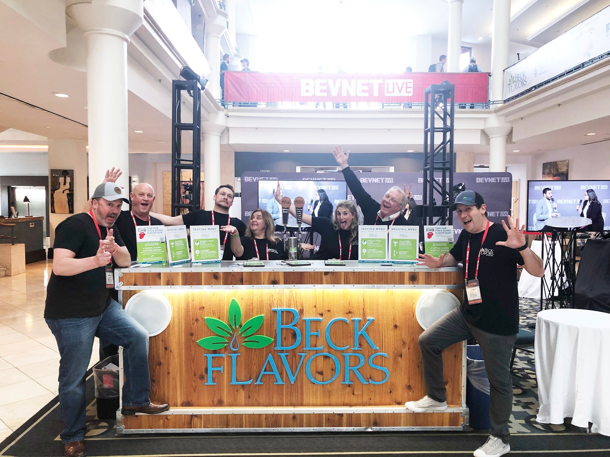 A group of exhibitors with raised arms pose behind a wooden counter with a Beck Flavors logo in a multi-story atrium.