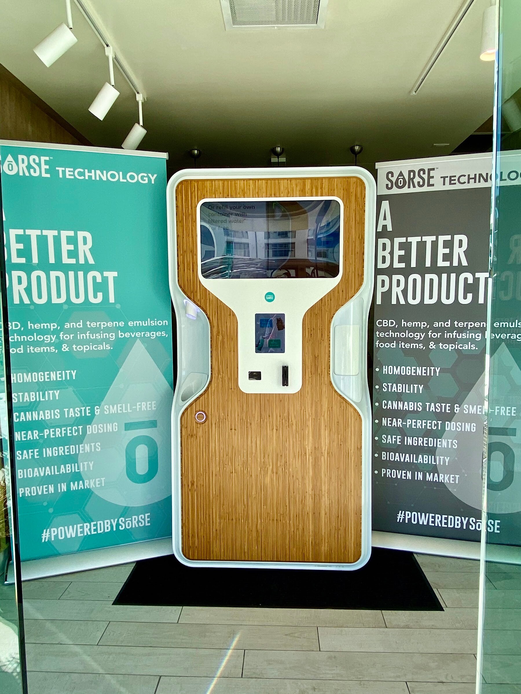 A tall white and wood dispensing unit with a screen sits between two teal Sorse Technology banners.