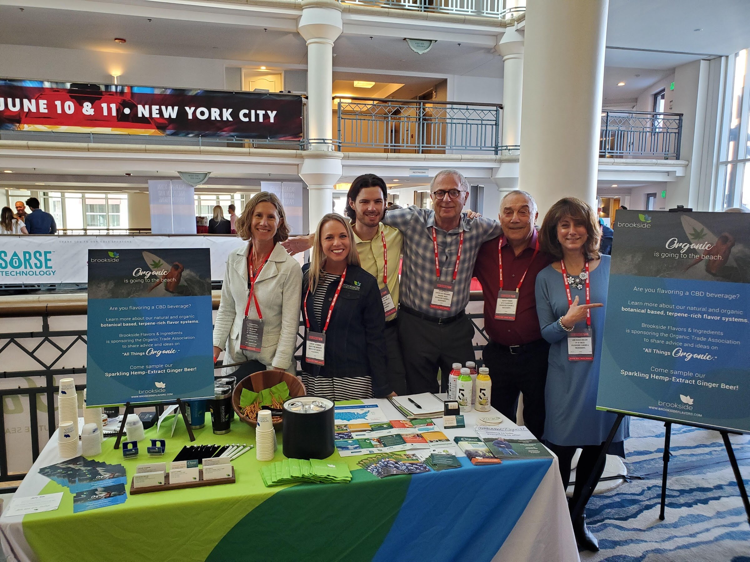 Seven people in conference badges stand behind a table covered with brochures and bottles under a brookside flavors banner.