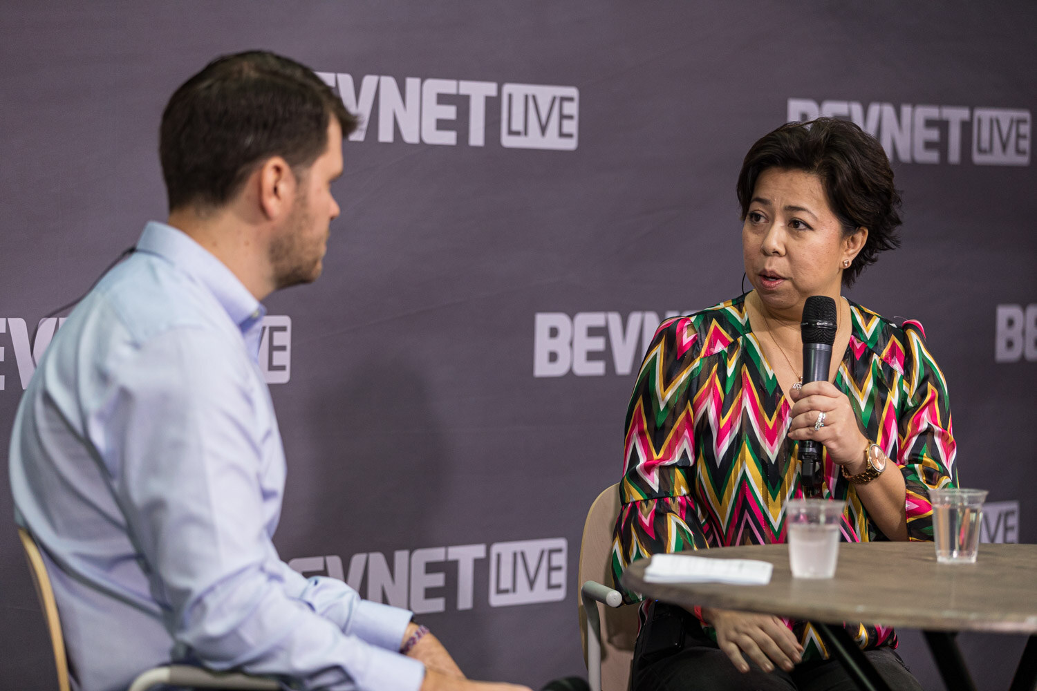 A man with his back turned and a woman in a zig-zag patterned blouse hold microphones at a round table.