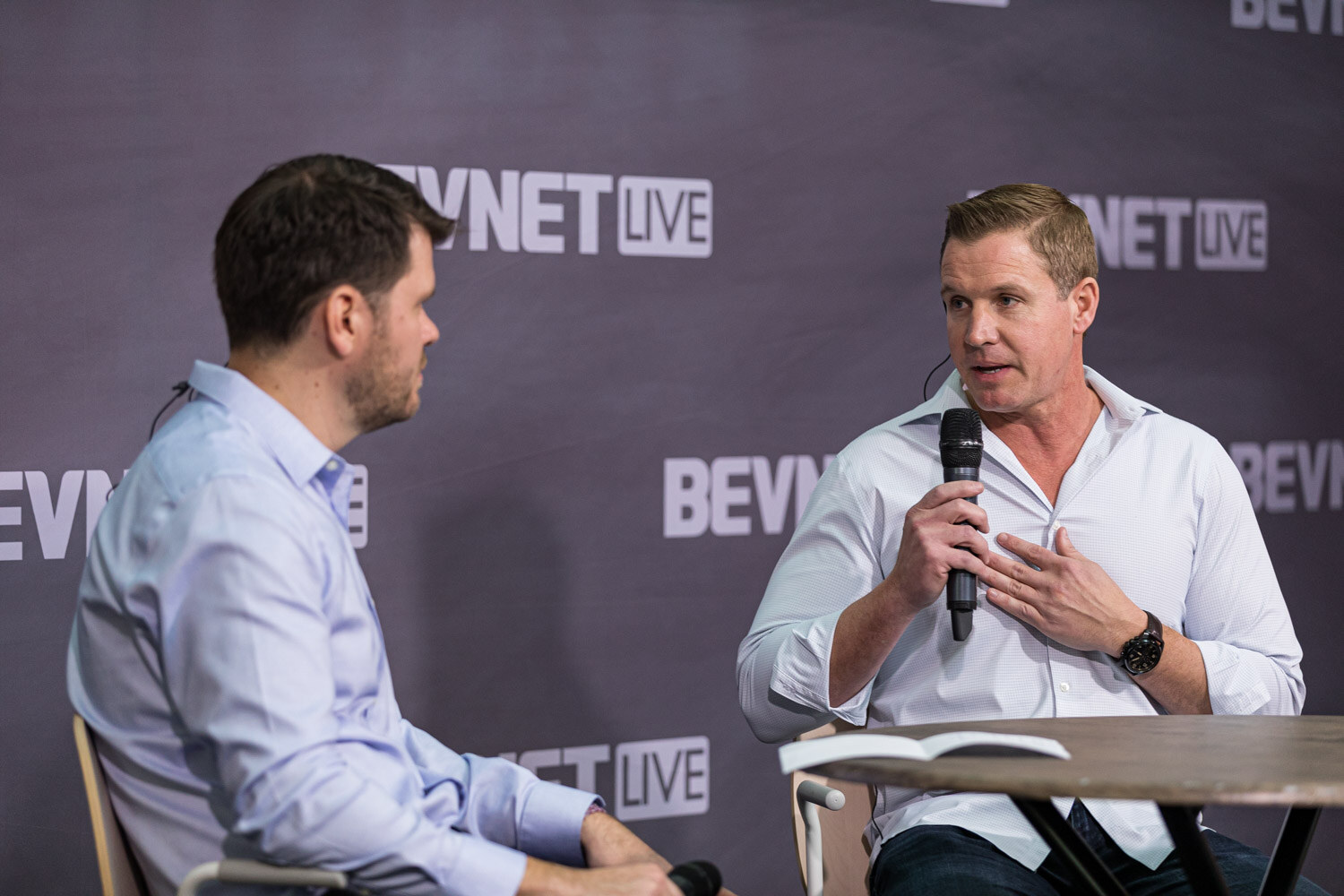 A man in a white shirt holds a microphone while a man in a light blue shirt listens.