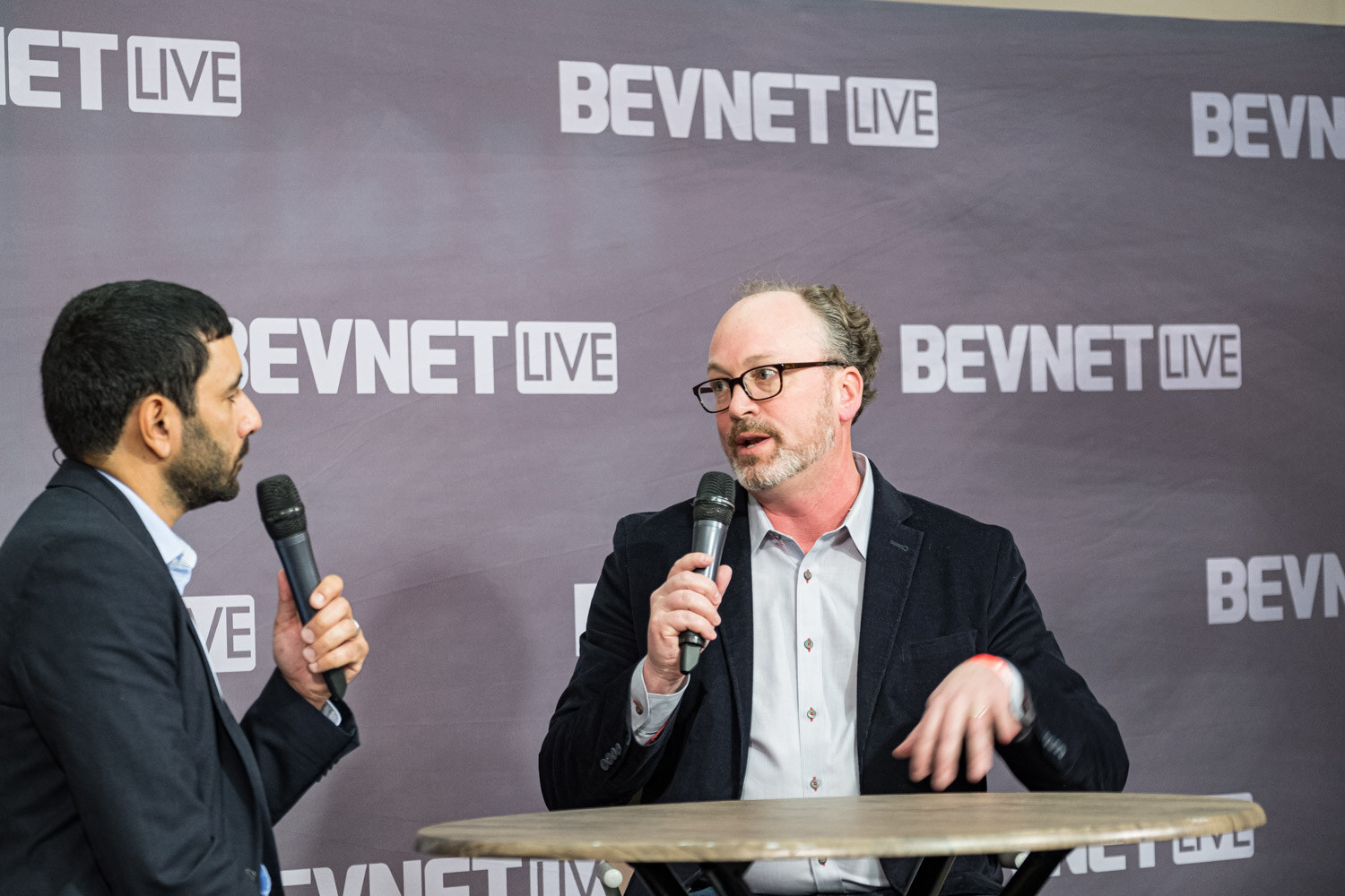 Two men with microphones sit at a small round table in front of a BevNET Live backdrop.