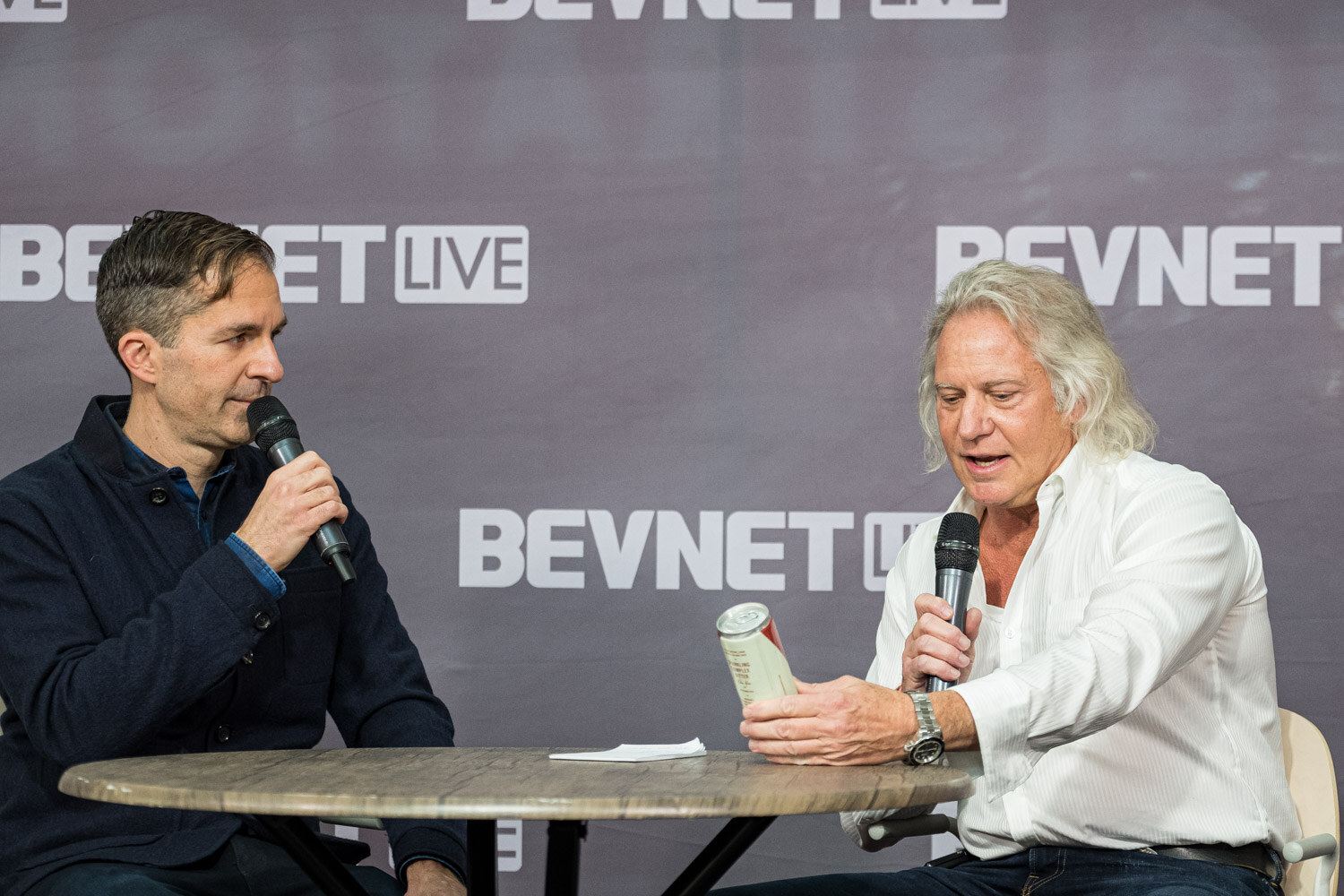 A man in a navy sweater and a man in a white shirt holding a can sit across a round table in front of a BevNET Live backdrop.