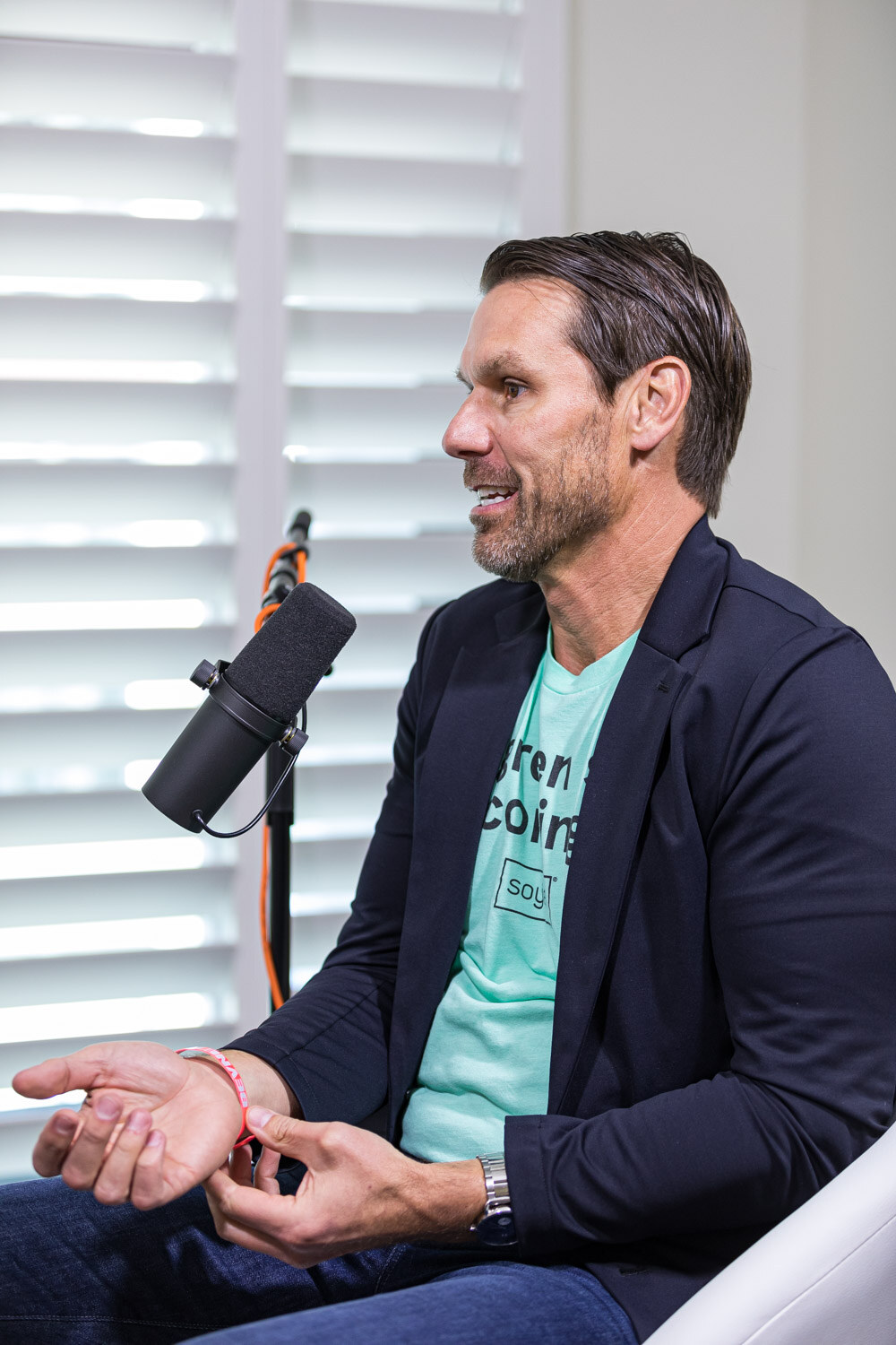 A man in a navy blazer and teal t-shirt gestures with both hands next to a microphone, with white window shutters behind.