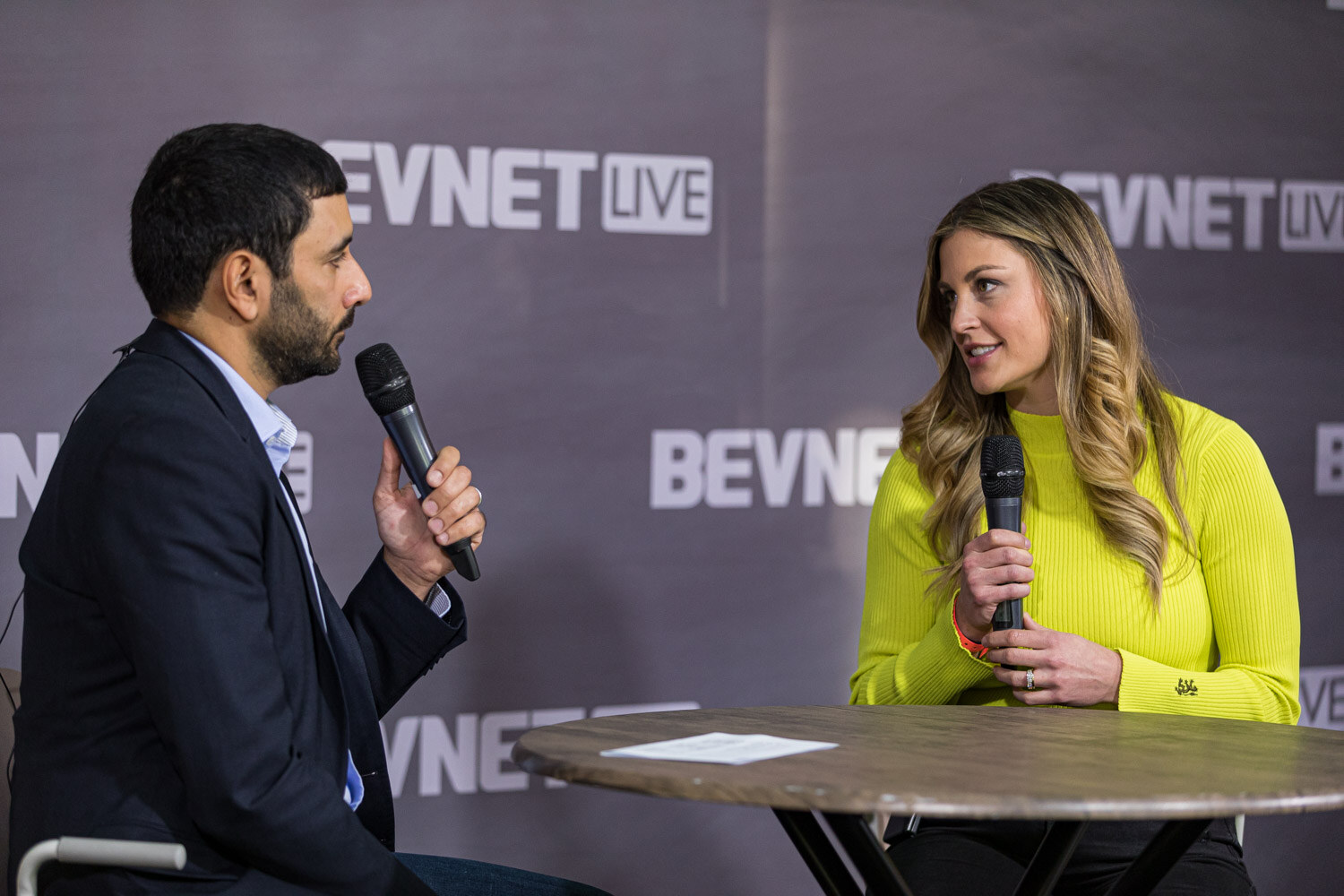 A woman in a bright yellow sweater holds a microphone and looks toward a man in a dark suit.
