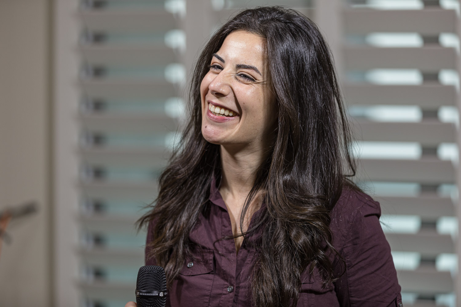 A smiling woman with long dark hair sits in front of white window shutters with a microphone in front of her.