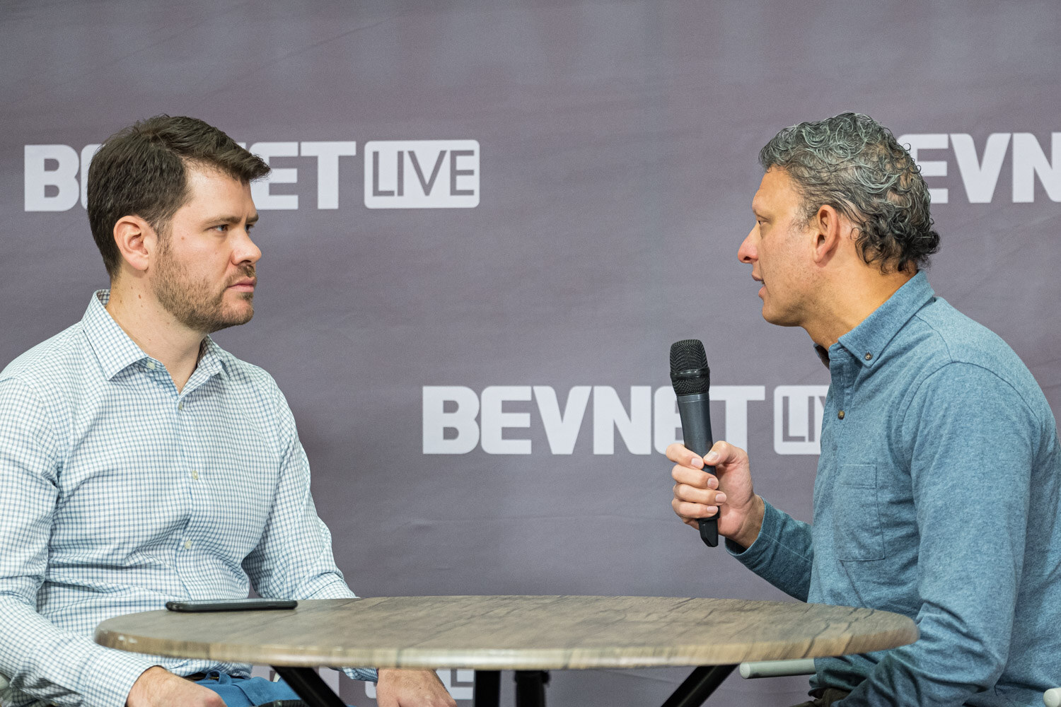 A man in a light blue checked shirt and a man in a blue shirt hold microphones at a small table in front of the BevNET Live backdrop.