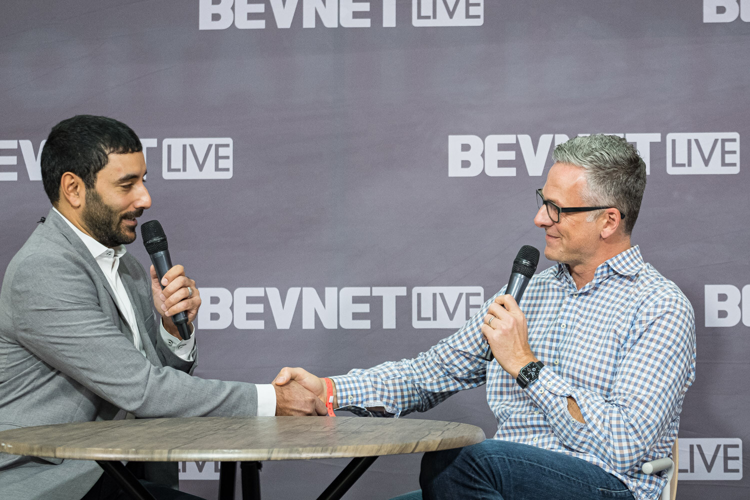 Two men with microphones shake hands over a small round table in front of a BevNET Live backdrop.