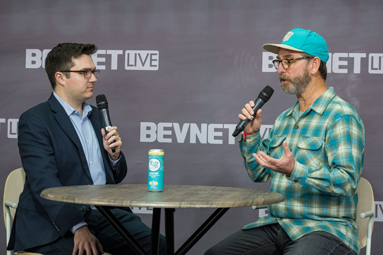 A man in a teal cap and plaid shirt talks with a man in a blue blazer next to a can on a round table.