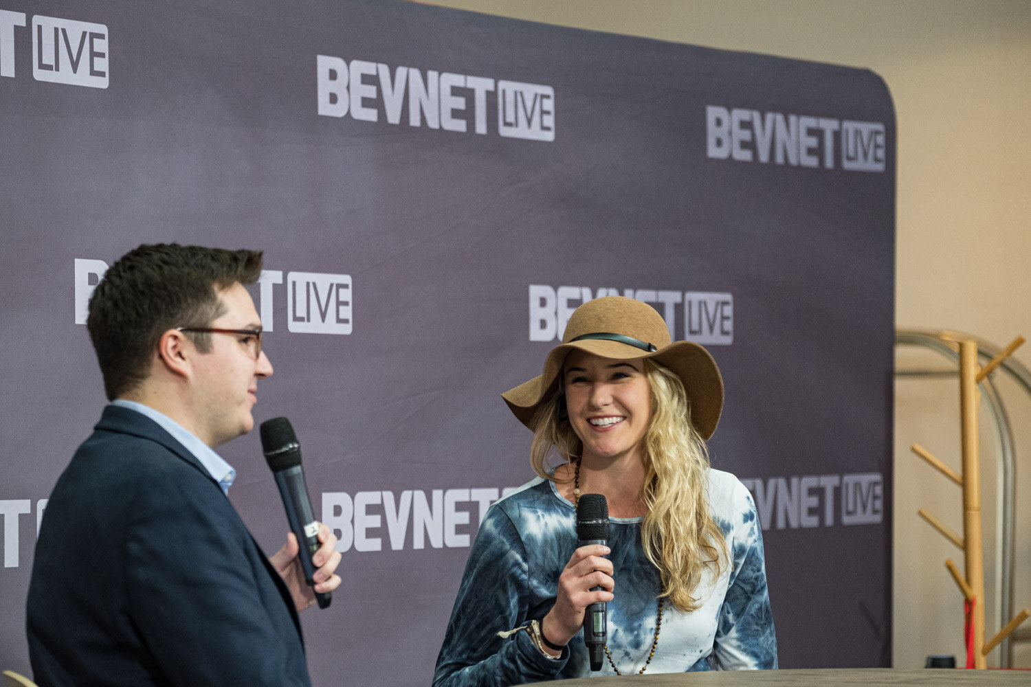 A woman in a brown hat and a tie-dye top holds a microphone while a man in a blue blazer listens.