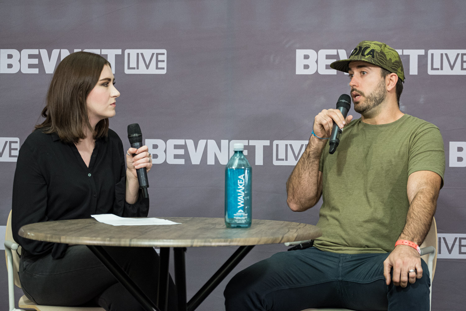 A woman in black and a man in a green shirt and camo cap sit at a round table with a water bottle in front of a BevNET Live backdrop.