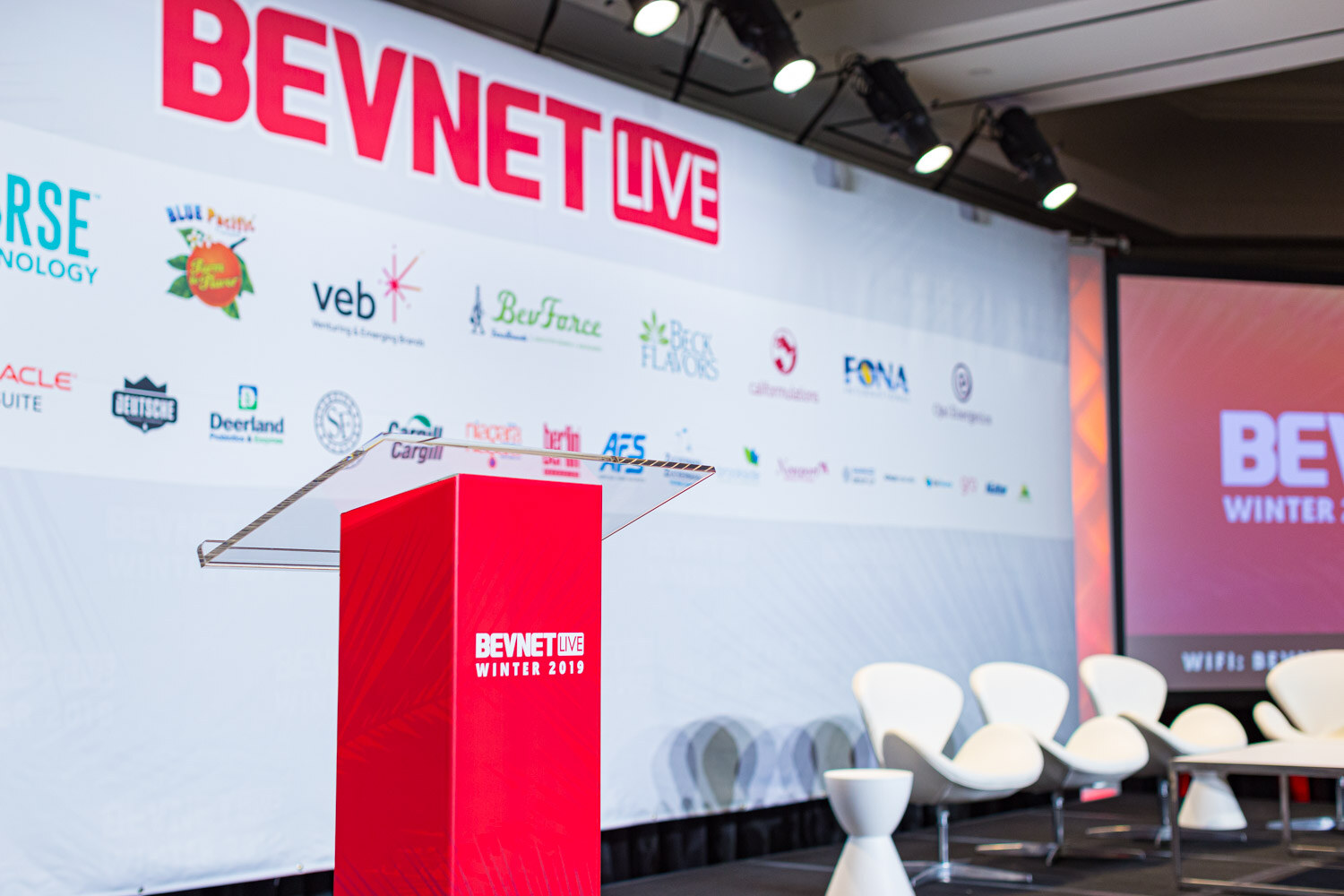 A red podium and white lounge chairs stand on stage in front of a BevNET Live backdrop with sponsor logos.