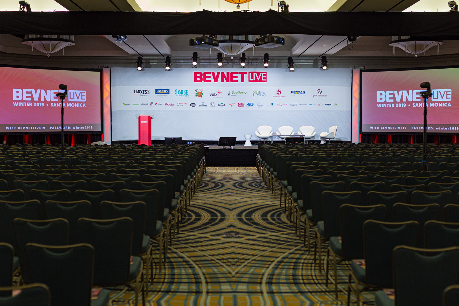 A wide ballroom with rows of empty chairs facing a stage with a BevNET Live backdrop and two side screens.
