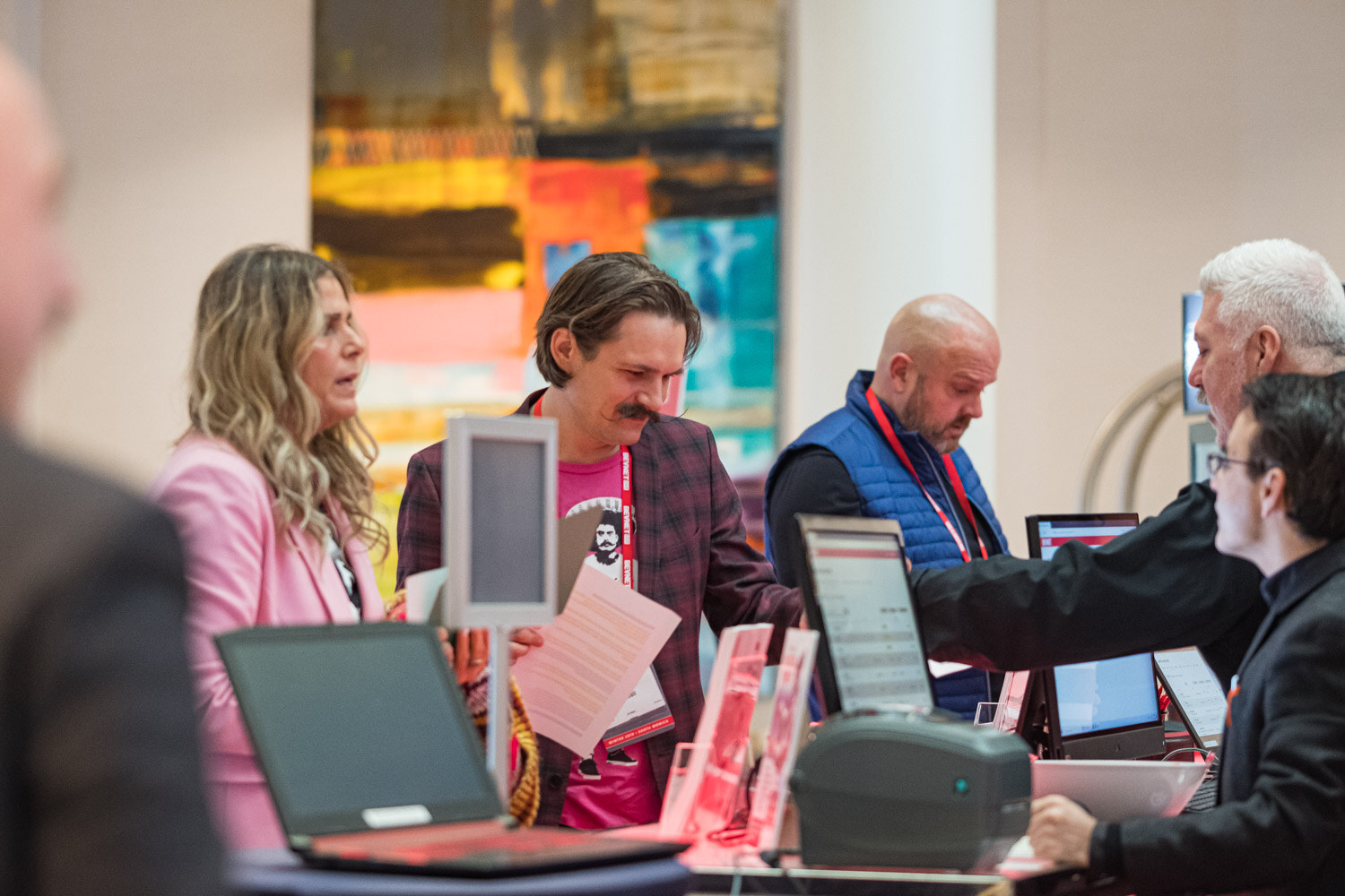 A woman in a pink jacket and a man in a plaid jacket with a pink shirt stand at a registration desk with laptops and a label printer.