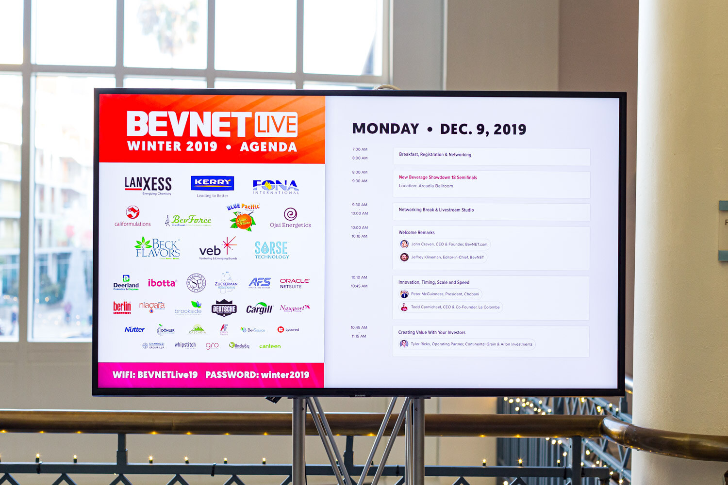 A wall-mounted screen titled BevNET Live Winter 2019 Agenda for Monday December 9, 2019 stands on a balcony.