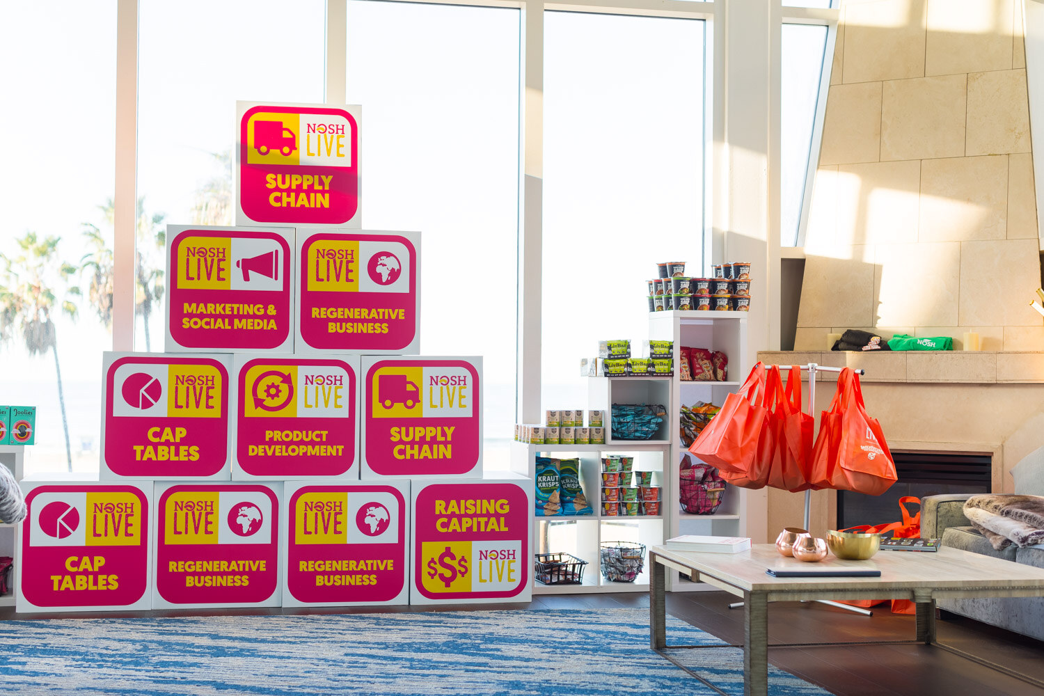 A pyramid of pink boxes marked Nosh Live with topics such as Cap Tables and Supply Chain stands beside shelves and a coffee table.
