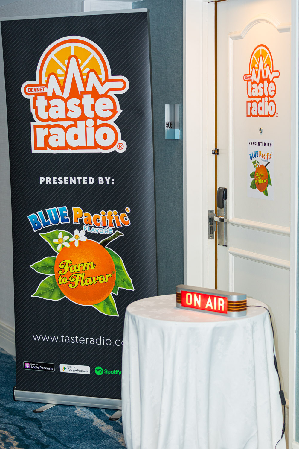 A tall Taste Radio banner stands beside a table with a glowing On Air sign next to a white door.