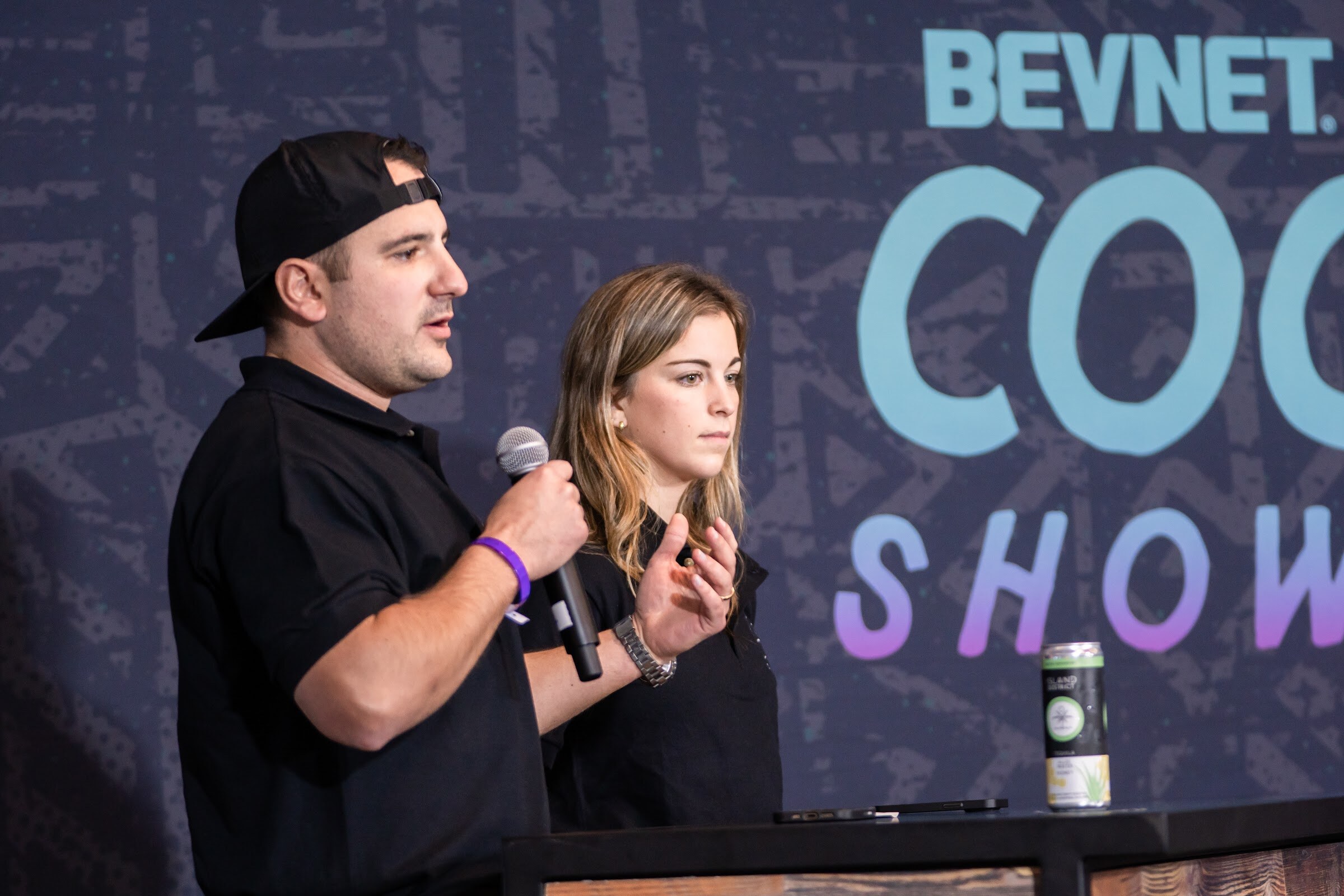 A man in a backwards black cap and a black Island District Company polo speaks into a microphone while a woman in the same shirt looks on.