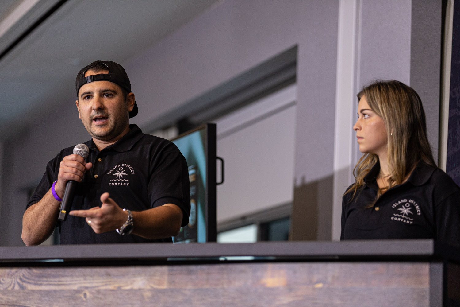 A man in a backwards black cap and Island District Company polo gestures while holding a microphone, beside a woman in the same polo, behind a wooden lectern.