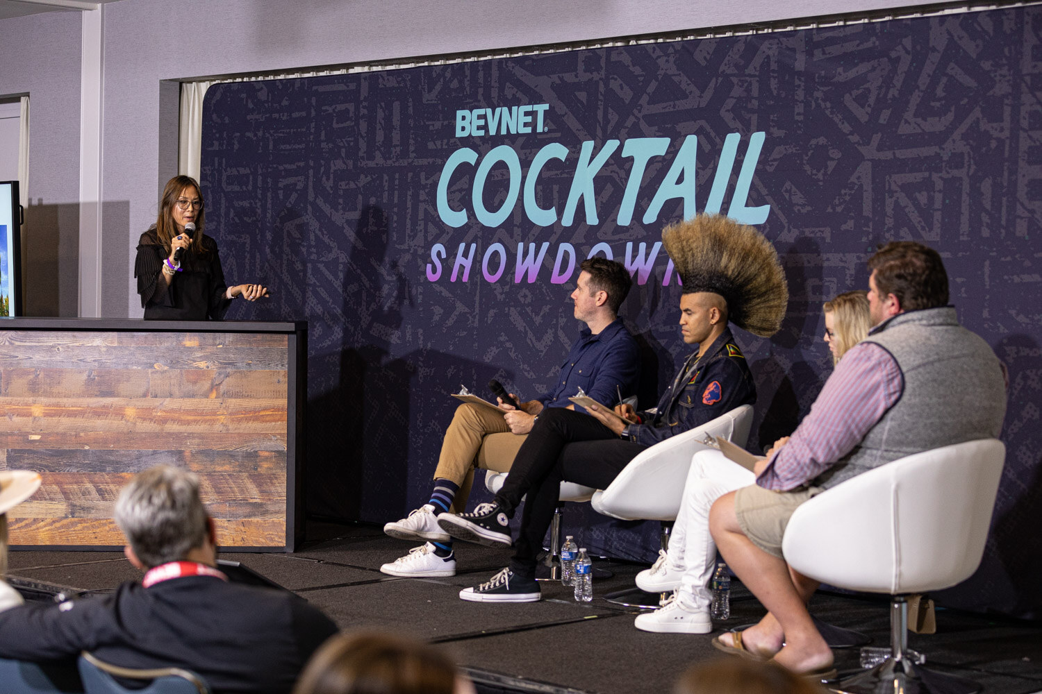 A woman at a wooden bar-style lectern speaks beside a Cocktail Showdown 2 backdrop with three judges seated in white chairs and an audience member in the foreground.