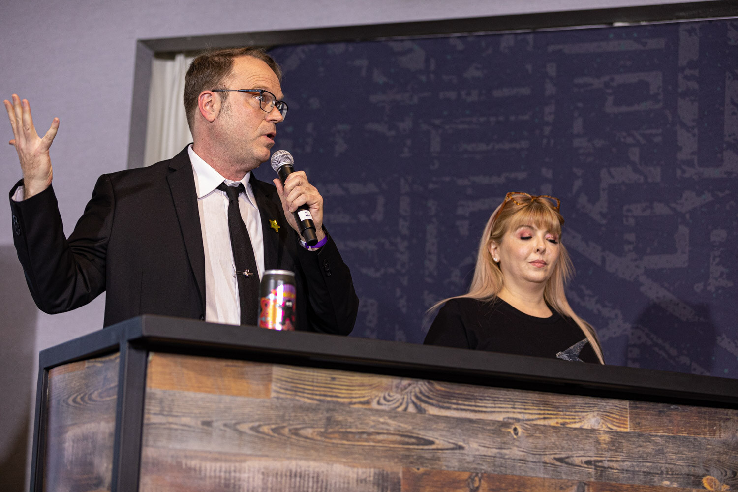 A man with glasses in a black suit and tie raises one hand while speaking into a microphone beside a blonde woman looking down, in front of a navy backdrop.