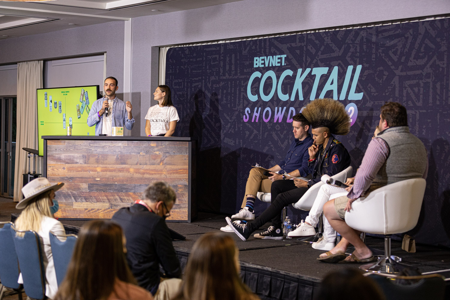 A wide view of a ballroom stage with a navy BevNET Cocktail Showdown backdrop, a green slide screen, two presenters at a wooden lectern and three judges seated in white chairs.