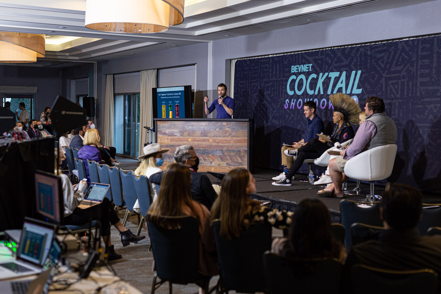 A wide view of a ballroom with an audience at tables and laptops in the foreground facing a stage with a Cocktail Showdown backdrop, a presenter at a lectern and judges seated.