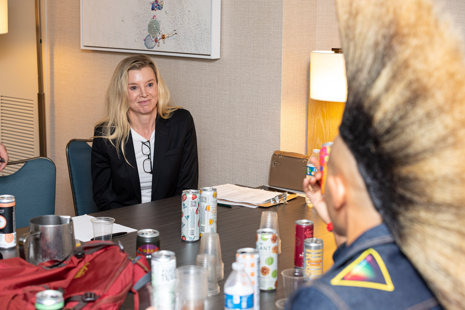 A blonde woman in a black blazer sits at a table with canned cocktails and cups, with the back of a judge's tall blond-tipped mohawk in the foreground.
