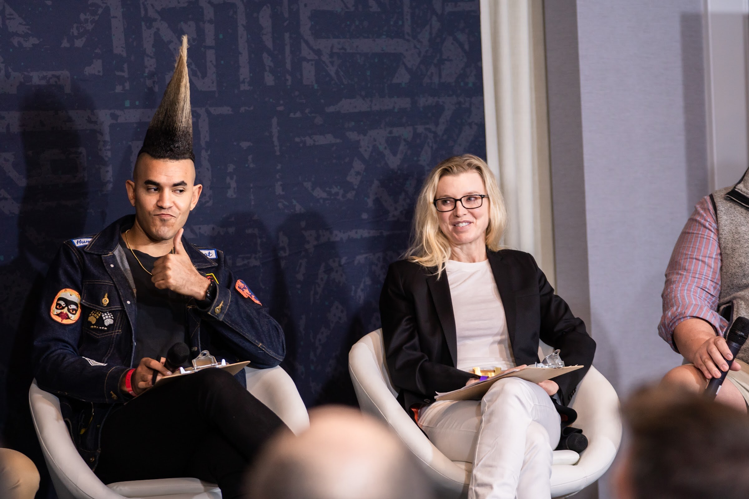A man with a tall mohawk and a denim jacket and a blonde woman in a black blazer sit in white chairs with clipboards in front of a navy backdrop.