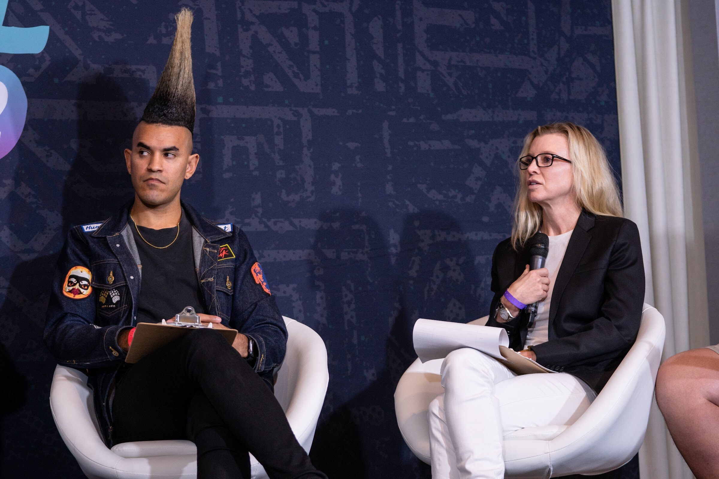 A blonde woman with glasses in a black blazer holds a microphone and papers while a man with a tall mohawk sits beside her.