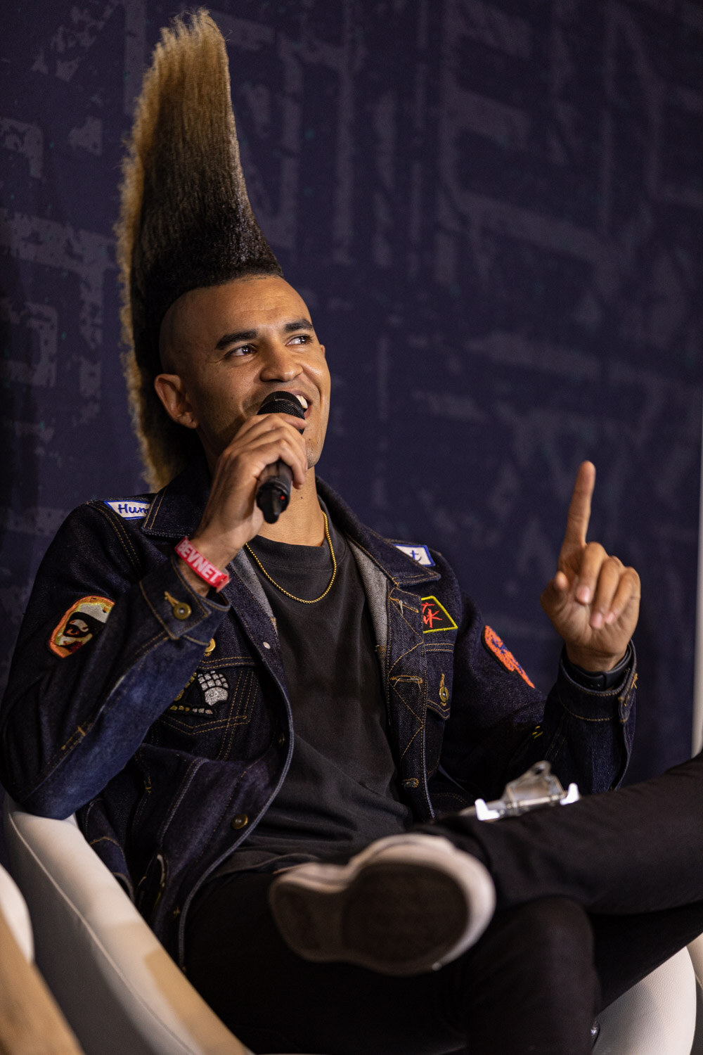 A man with a tall blond-tipped mohawk in a denim jacket holds a microphone in one hand and points up with the other while seated.