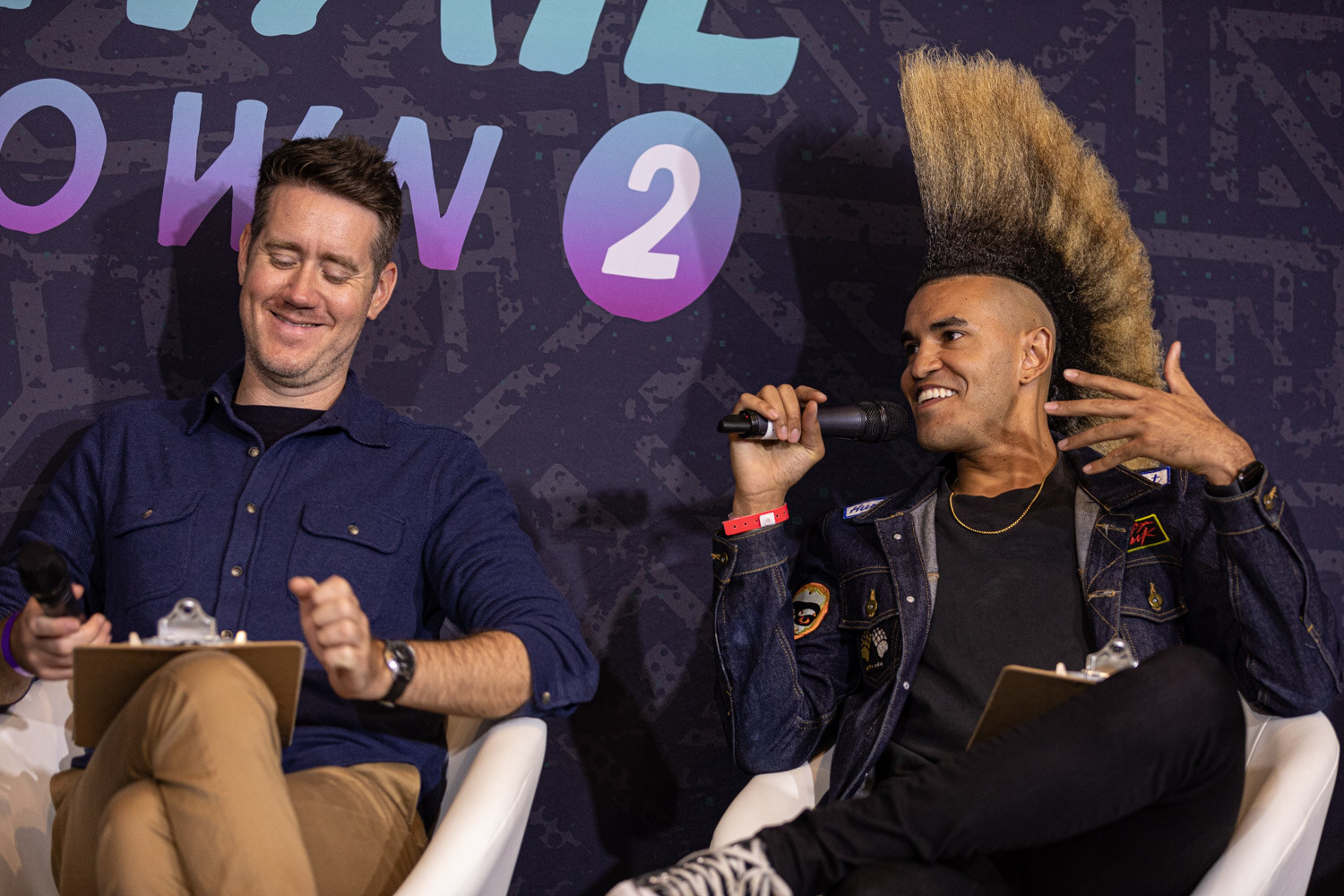 A man in a navy shirt looks down laughing with a clipboard on his lap next to a man with a tall mohawk who speaks into a microphone with a raised hand.