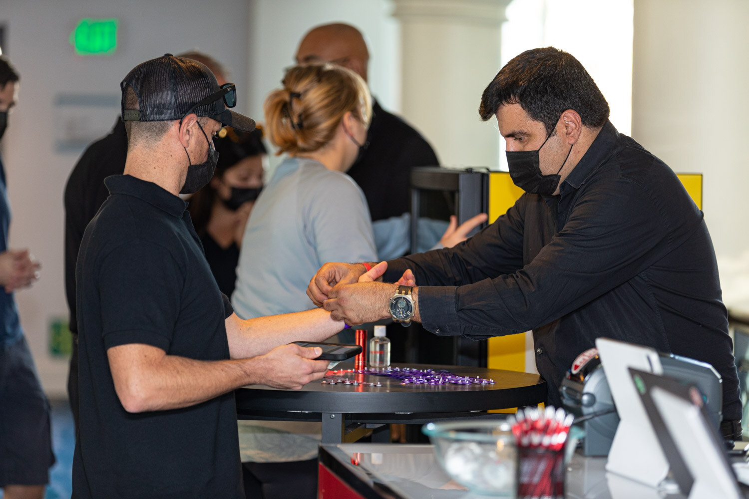 A man in a black shirt and mask fastens a wristband on another man's wrist at a registration table with purple wristbands.