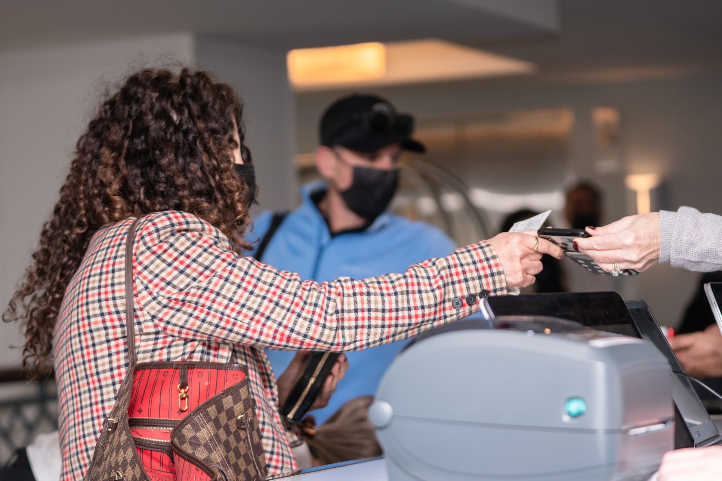 A woman with long curly hair in a mask hands a card across a desk with a label printer to a staffer in a blue shirt and black cap.