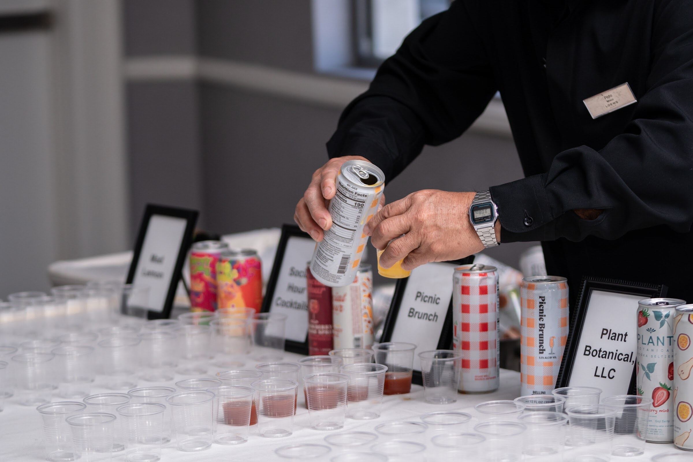 A hand holds a can above a white table of plastic sample cups and cans next to placards reading Mad Lemon, Social Hour Cocktails, Picnic Brunch and Plant Botanical LLC.