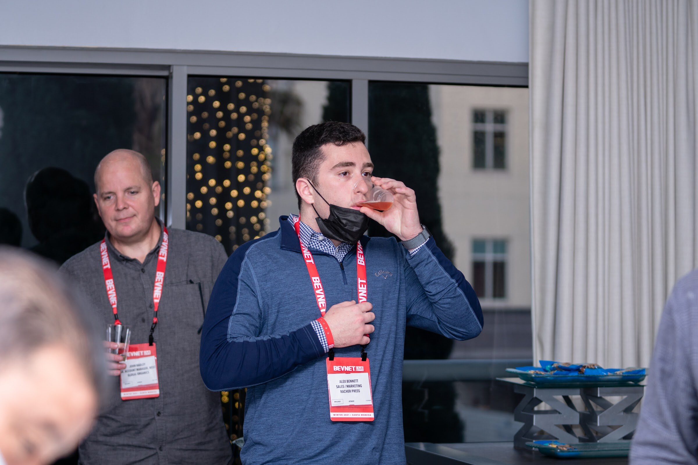 A man in a navy quarter-zip with a BevNET lanyard sips from a small cup, with a man in a gray shirt and BevNET lanyard standing behind him.