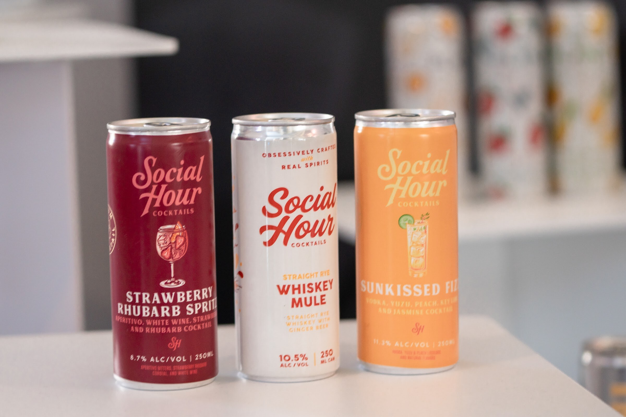 Three cans labeled Social Hour Cocktails in red, white and orange on a white table with more cans blurred behind.