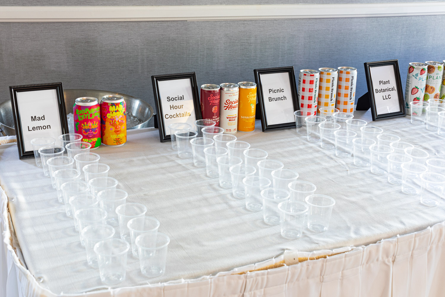 A long white table with rows of clear plastic cups and cans behind placards reading Mad Lemon, Social Hour Cocktails, Picnic Brunch and Plant Botanical LLC.