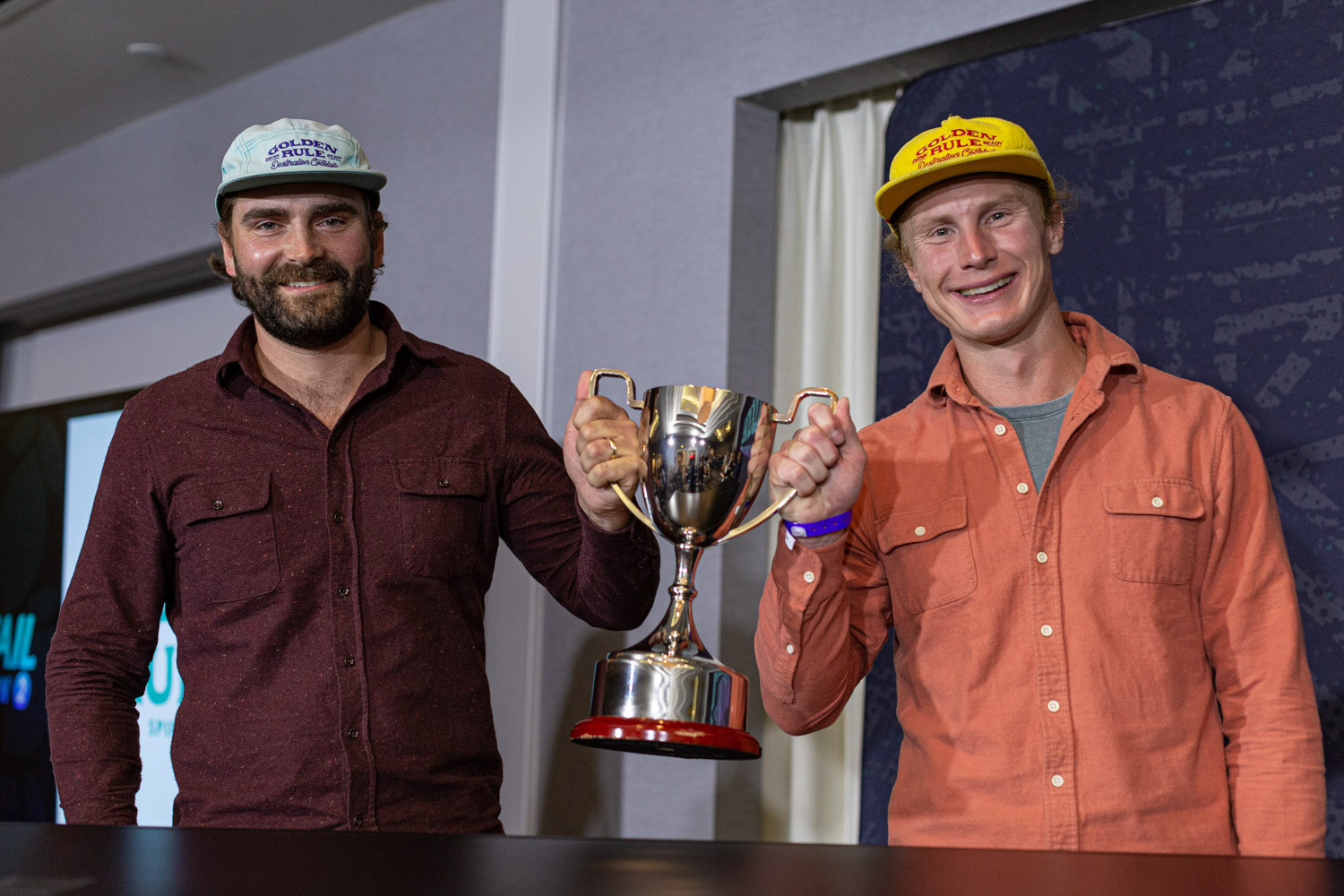 Two smiling bearded men in caps and button-up shirts, one in maroon and one in orange, hold a silver trophy cup together.