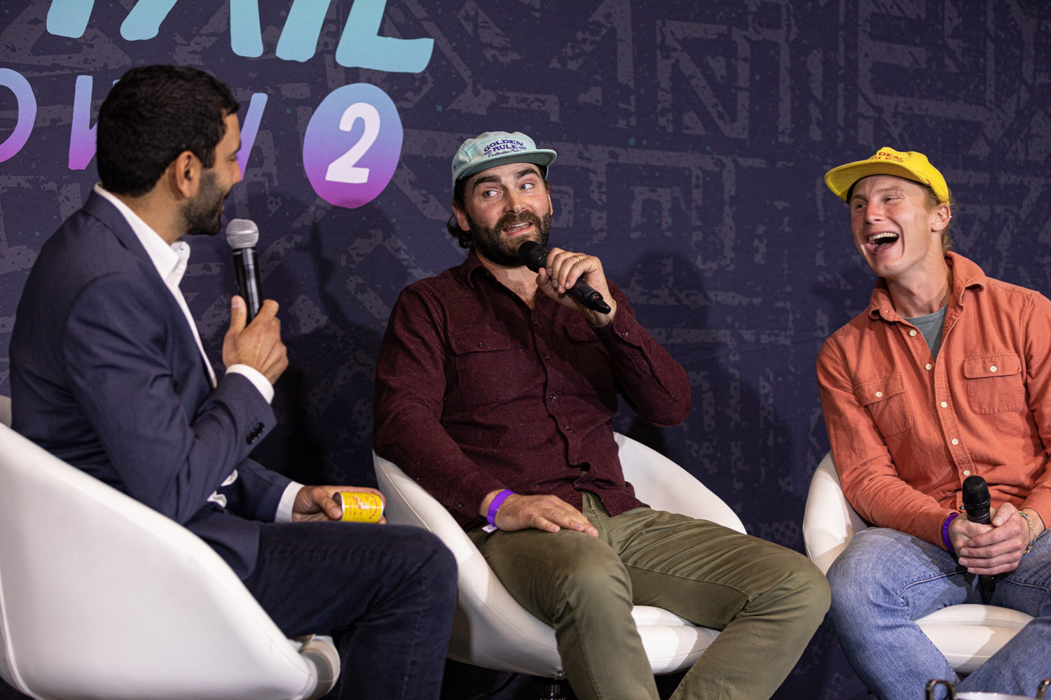 A man in a navy blazer holds a microphone toward a man in a maroon shirt and cap and a laughing man in an orange shirt and yellow cap in front of a Cocktail Showdown 2 backdrop.