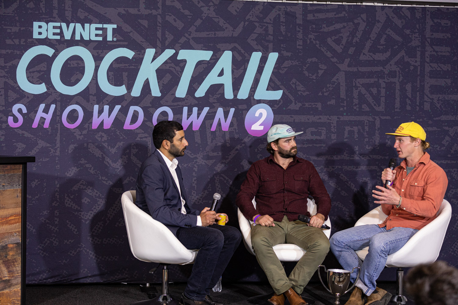 Three men in white chairs in front of a navy BevNET Cocktail Showdown 2 backdrop, with a silver trophy cup on the floor beside them.