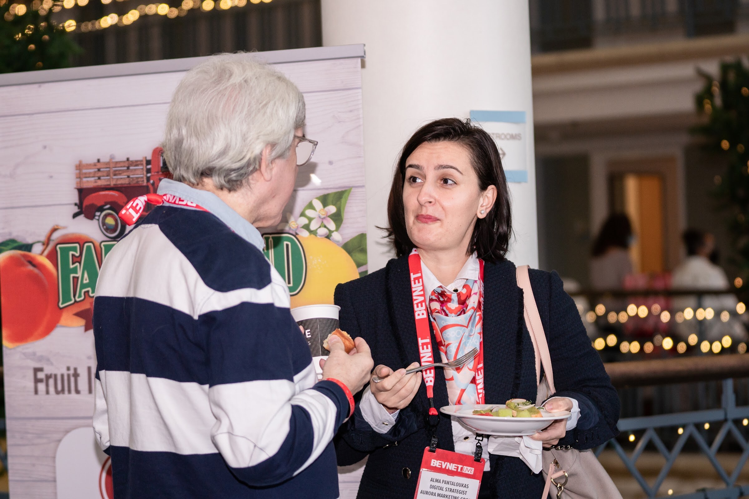 A woman with a BevNET lanyard holds a plate of food while a man in a striped sweater faces her in front of a fruit-themed banner.