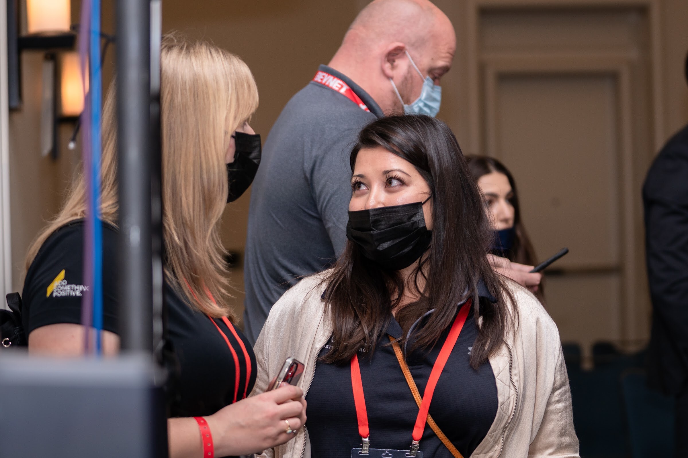A woman with dark hair in a face mask and a red lanyard looks up toward a man in a gray shirt and mask, with others beside them.