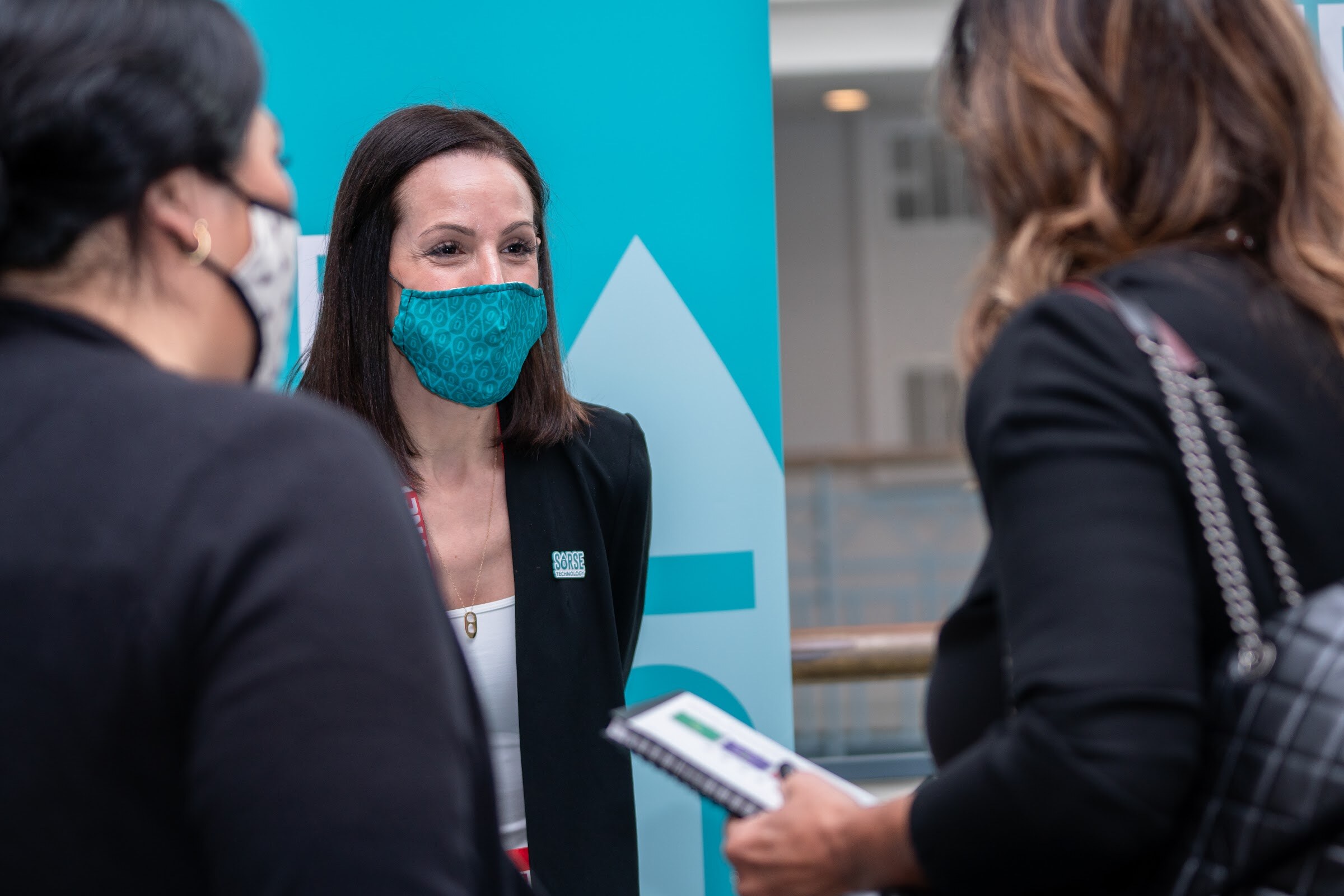 A woman in a black blazer and a patterned teal face mask stands in front of a teal banner, with two attendees in the foreground.