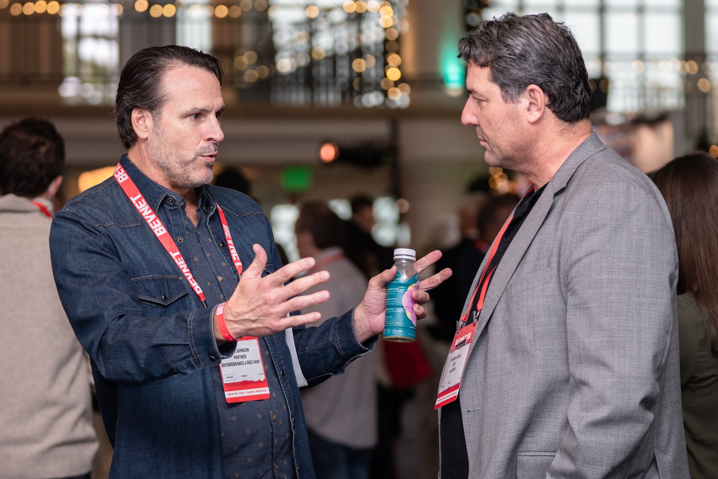 A man in a denim shirt with a BevNET lanyard gestures while holding a small bottle and talks with a man in a gray blazer.