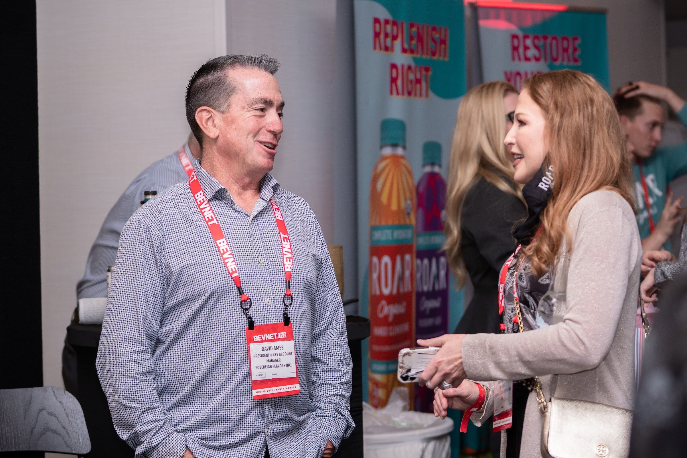 A man in a gray patterned shirt and a red BevNET lanyard smiles at a woman with long auburn hair in front of teal Roar Organic banners.